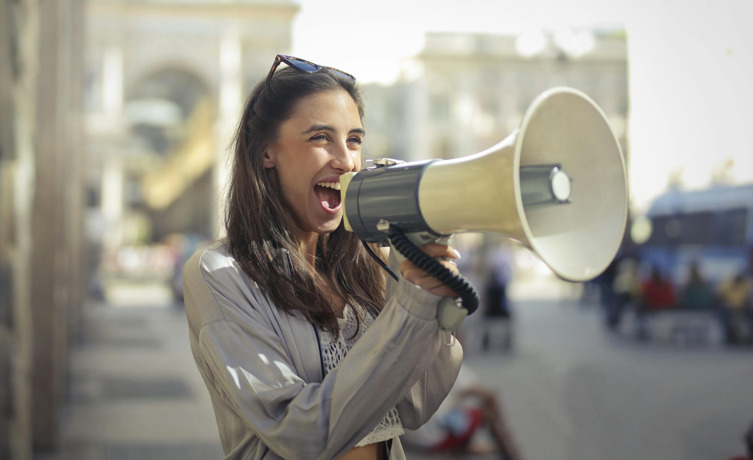 Cheerful young woman in a casual outfit shouting into a megaphone on a sunny day.